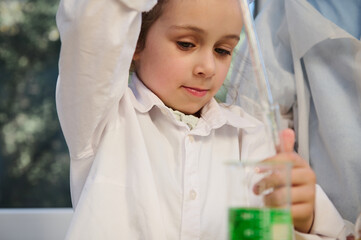 Beautiful mischievous child girl holding a glass pipette, and dripping few drops of green chemical solution from a flask into a test tube during a chemistry lesson, in the school science laboratory