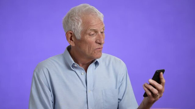 Happy fun elderly man 70s in blue shirt gets video call using mobile cell phone talk wave with hand isolated on plain light purple background studio portrait 