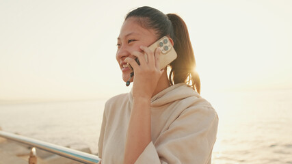 Smiling Asian girl in sportswear talking on the phone while standing on the embankment on modern buildings background in the morning light