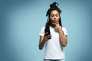 Curly haired ethnic woman uses mobile phone checks messages holds modern cellular in hands isolated over blue background