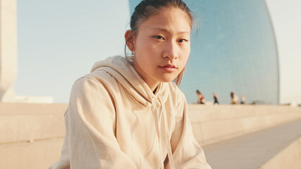 Asian girl in sportswear resting after workout while sitting on steps on modern buildings background at sunrise. Young woman turns her head and looks at the camera.