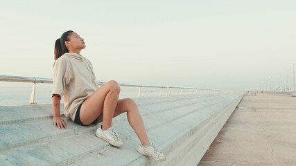 Asian girl wearing sportswear sits on the waterfront, enjoys the fresh air at morning time