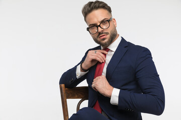arrogant young businessman sitting on chair and adjusting red tie