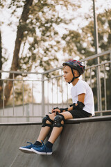 Cute kid boy child in a helmet sitting in a special area in skatepark and holding skateboard. Summer sport activity concept. Happy childhood.