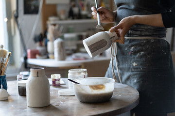 Ceramist wearing black apron for work paints clay vase. Craftmaster prepares necessary tools for...