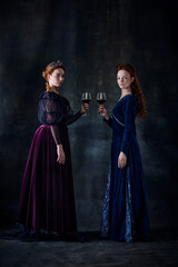 Portrait of two beautiful women in image of queens with wine glasses isolated over dark background
