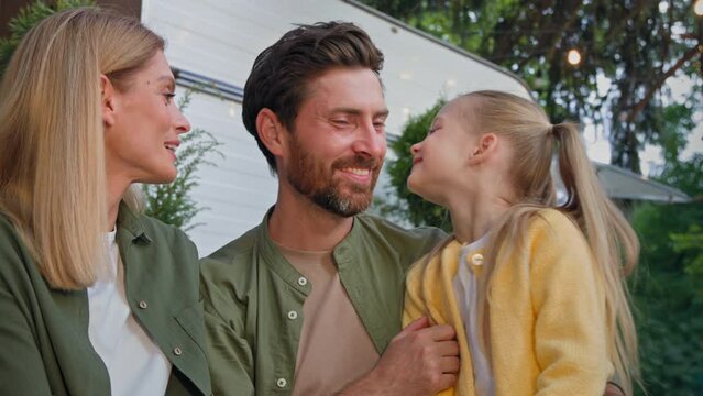 Caucasian happy loving family talking outdoors dad touching nose of little daughter child girl and mother wife kissing bearded father on cheek hugging love relationship affectionate cuddling together