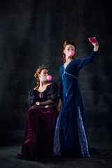Portrait of two beautiful women, royal persons taking selfie with bubblegum isolated over dark...