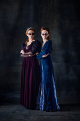 Portrait of two beautiful women in image of queen and princess in stylish sunglasses isolated over dark background