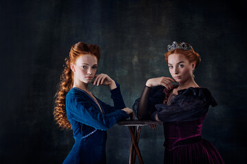 Portrait of two beautiful women in image of queen and princess isolated over dark background. Royal...