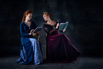 Portrait of two beautiful women in image of queens reading isolated over dark background. Evening...