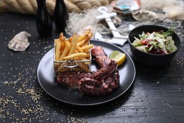 Grilled octopus tentacles served with potato fries in metal serving basket, on a black plate. Lunch set in a restaurant.