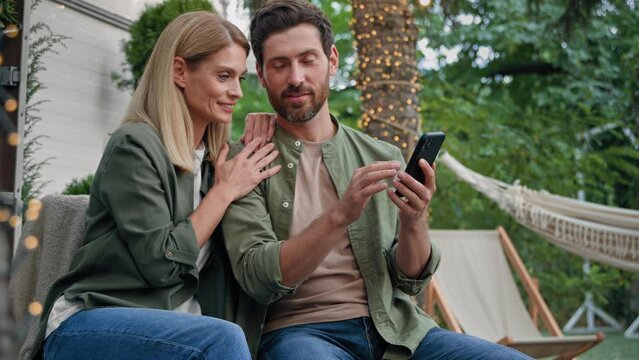 Married couple husband and wife Caucasian bearded man and blond woman 40s spouses outdoor in backyard at campsite looking at phone choose buy online booking with smartphone using mobile e-shopping app