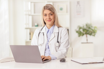 Young female doctor sitting in an office with a laptop computer, typing and smiling