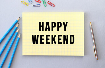 HAPPY WEEKEND text in notebook on gray background next to pencils, pen and paper clips.