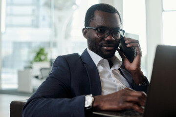 Young busy ceo or employee in formalwear speaking by mobile phone and looking at laptop screen while working over new project