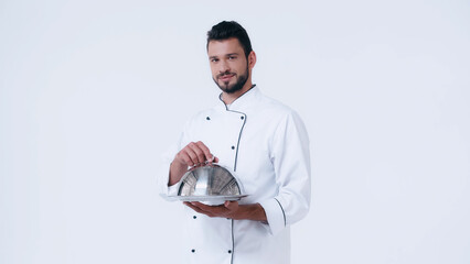 brunette man in chef jacket holding serving dish with cloche and looking at camera isolated on white.