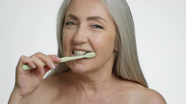 Dental care in senior age. Positive elderly woman brushing her teeth with natural brush, white studio background