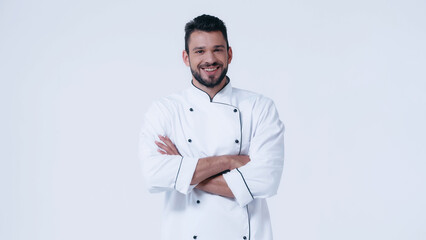 young and happy man in chef jacket standing with crossed arms isolated on white.