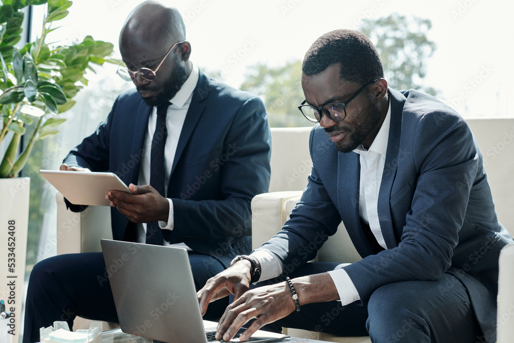 Poster Young broker in elegant suit pressing keys of laptop keypad during network or while analyzing online financial data against colleague
