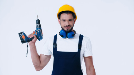 Fototapeta na wymiar positive builder in hardhat and overalls holding electric drill and looking at camera isolated on white.
