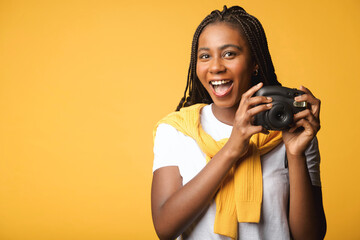 Cheerful african-american woman holding polaroid camera standing isolated on yellow. Tourist woman...