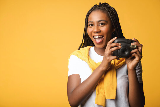 Cheerful african-american woman holding polaroid camera standing isolated on yellow. Tourist woman making instant photo with retro camera. Photographer concept