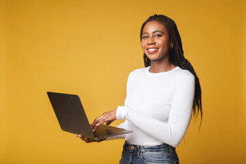 Attractive woman working on new post for her job on laptop, looking at camera with happy inspired smile, watching video on internet. Indoor studio shot isolated on yellow background