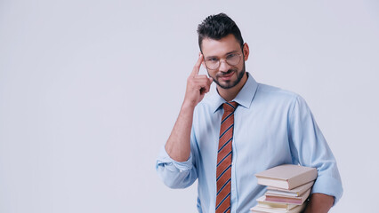 smiling teacher holding pile of books and pointing at head isolated on grey.