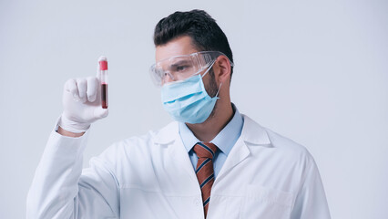 immunologist in medical mask and goggles looking at test tube with blood sample isolated on grey.