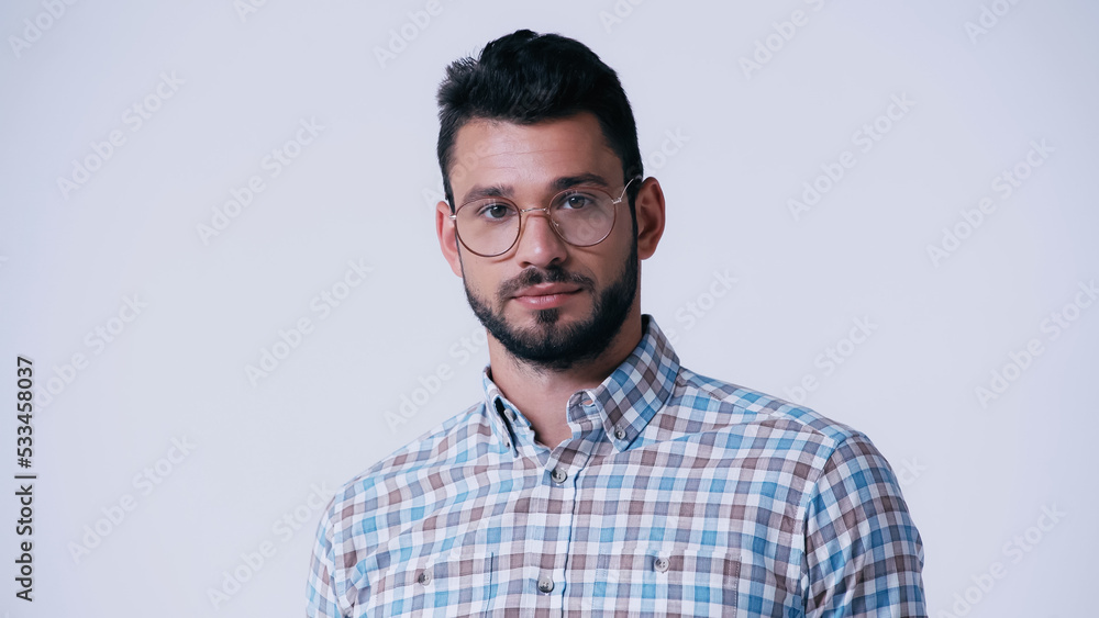 Wall mural young bearded nerd in eyeglasses and plaid shirt looking at camera isolated on grey.