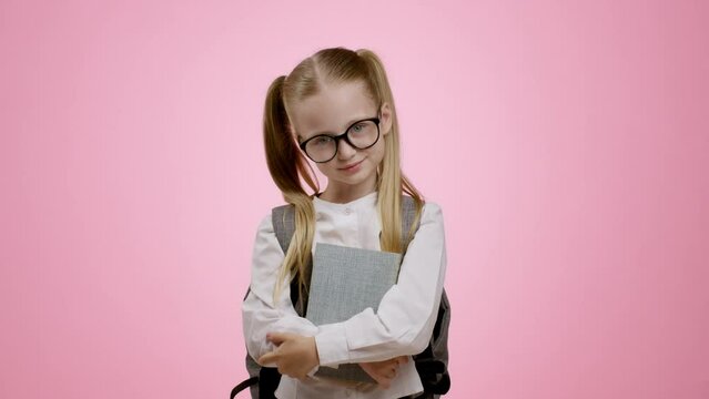 Little genius. Cute pretty little schoolgirl carrying backpack and books, fixing her eyeglasses and smiling to camera