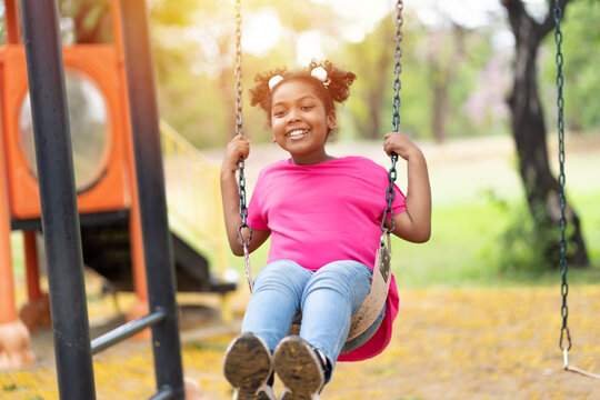 Smiling African American Child Girl Playing On Swing At The Playground. Happy Girl Having Fun On Swing Outdoor