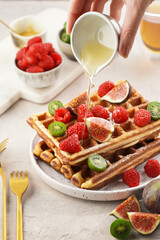 Pouring honey over fresh homemade sweet belgian waffles with powdered sugar with fresh raspberries, kiwi berries and figs on white plate