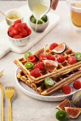 Pouring honey over fresh homemade sweet belgian waffles with powdered sugar with fresh raspberries, kiwi berries and figs on white plate