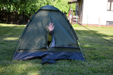 Child hand waving hello or hi from closed small tent on lawn green grass in backyard. Outdoor recreation, camping concept.
