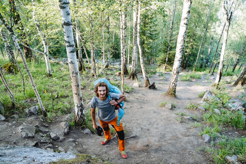 A tourist climbs the mountains with a heavy backpack, a mountain trail, a guy looks into the camera, a satisfied face, a smile, a hike in the mountains, hiking alone.