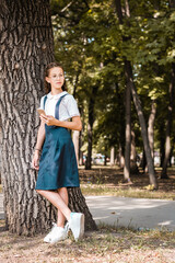 A pupil in eye glasses with a smartphone in her hand stands near a tree in the park. Vertical view