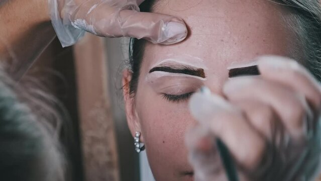 Eyebrow tinting procedure with henna in a beauty salon. The master brow corrects the shape of the eyebrows with a brush. A professional cosmetologist makes make-up. Face of a young girl close-up. 4K