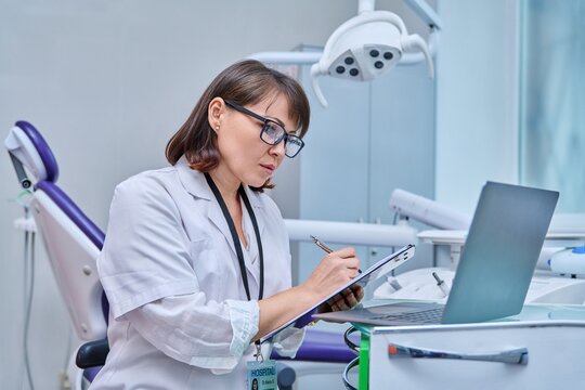 Doctor dentist working in office, using laptop, making notes on clipboard