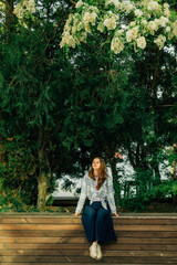 a young woman sits on a wooden bench in summer with a smile on her face in the shade of a large tree