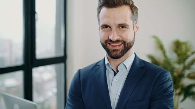 Video portrait of handsome confident elegant successful caucasian bearded man, in a suit, top manager, company ceo, financial director, stand in the office, arms crossed, looks at the camera, smiling