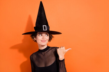 Photo portrait of charming young girl offer look empty space promo dressed trendy black halloween witch look isolated on orange background