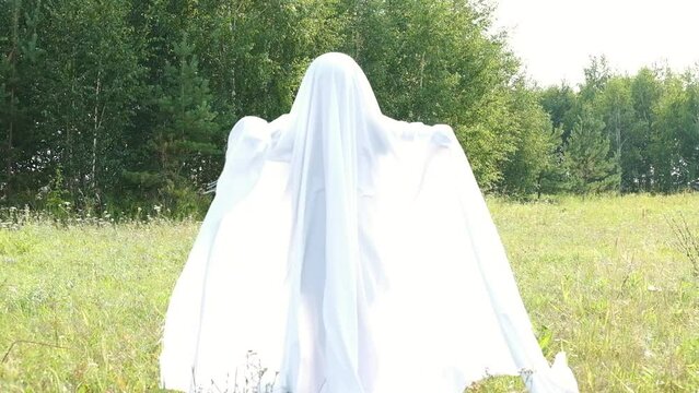 Funny Halloween concept. A small child dressed in a white cloth scares everyone on the street. The ghost is dancing. A child in a carnival costume of a ghost in the garden. trick or treat