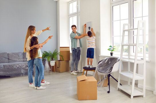 Happy family who recently bought house are decorating their new home together. Parents and children unpacking belongings from cardboard boxes and choosing place on wall in living room to hang picture