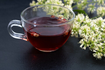Mug with pink apple tea on a blue cloth on a black background with guaco flowers on the side. Mention of treatment with alternative, herbal and natural medicine.