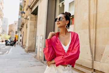 Attractive fashionable girl posing on summer old european street and looking aside. Stylish young woman with dark hair wearing pink shirt and and white shorts