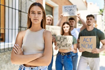 Young hispanic activist woman with arms crossed gesture standing with a group of protesters holding...