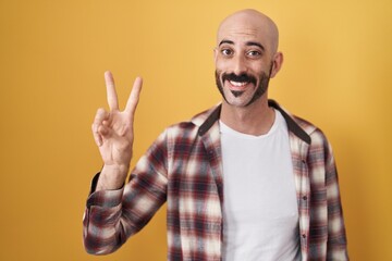 Naklejka premium Hispanic man with beard standing over yellow background showing and pointing up with fingers number two while smiling confident and happy.