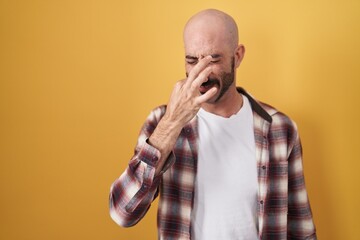 Hispanic man with beard standing over yellow background smelling something stinky and disgusting,...